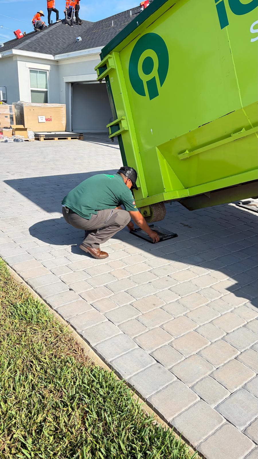 iCdumpsters technician installing Rolliskate driveway protection pads under a green dumpster on a residential paver driveway before roofing debris drop-off